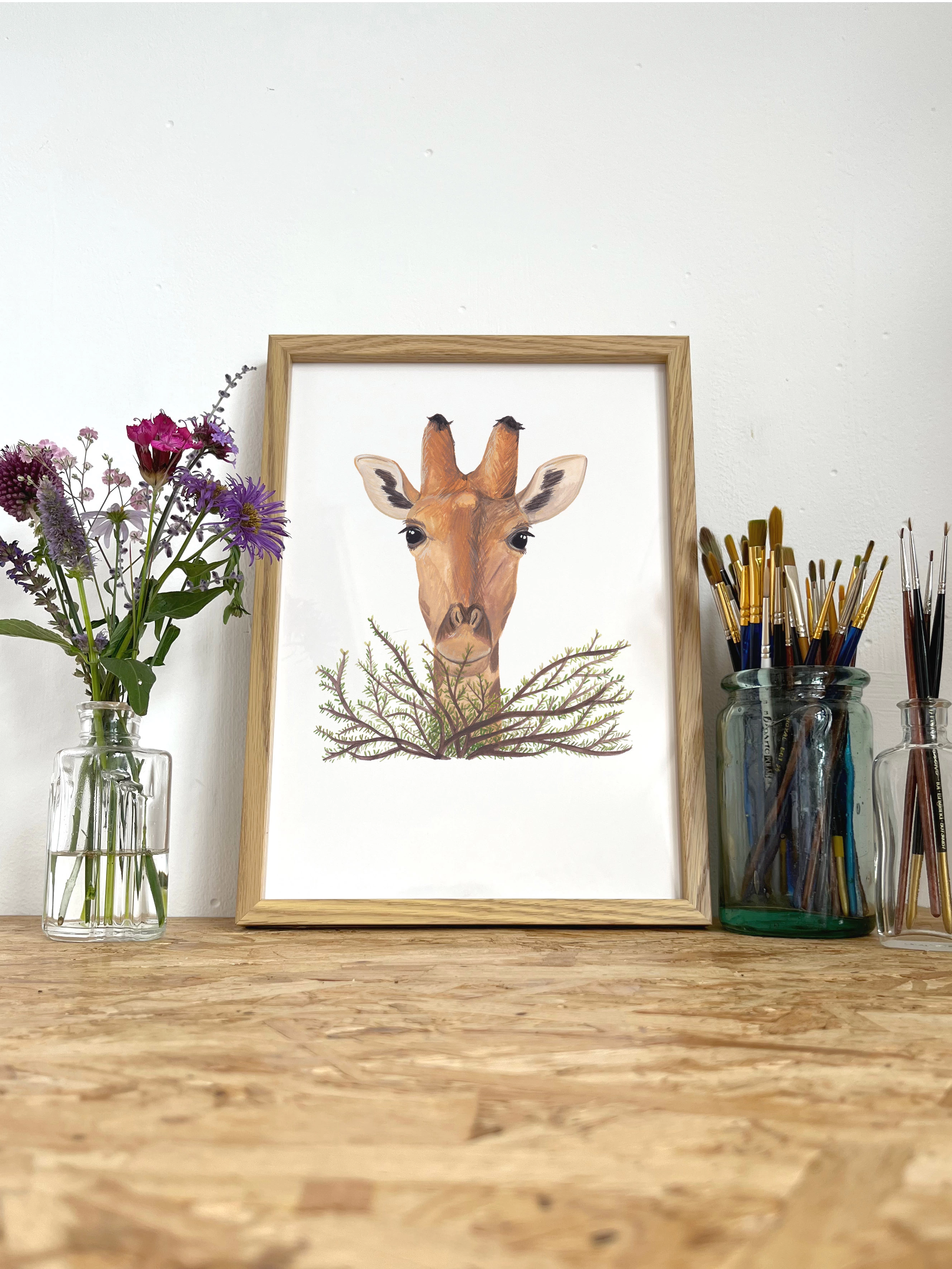 A print of anm illustration of a giraffe's head popping out over a bush, in a frame next to some flowers and a glass pot of paint brushes