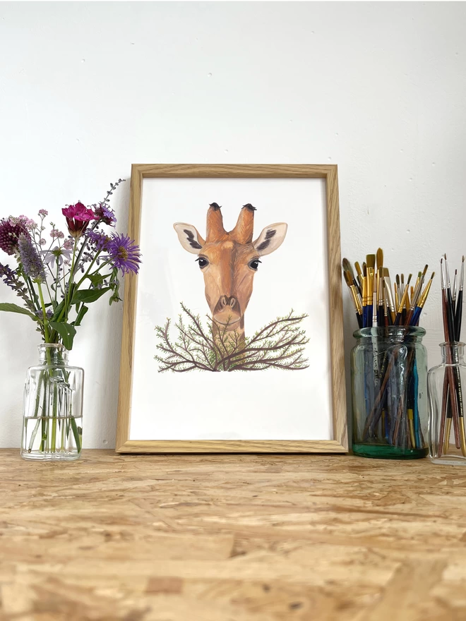 A print of anm illustration of a giraffe's head popping out over a bush, in a frame next to some flowers and a glass pot of paint brushes