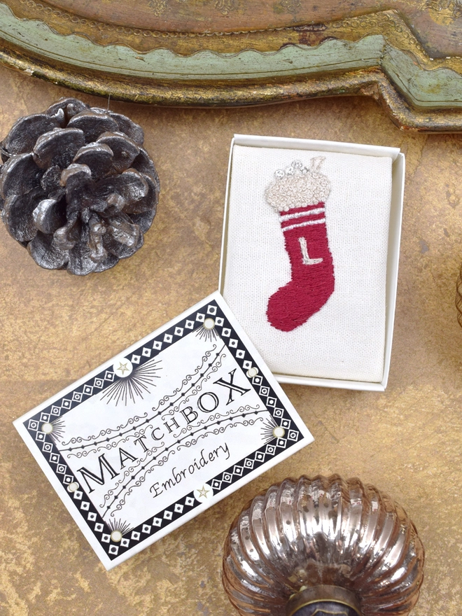 An embroidered stocking, inside a Matchbox displayed on a gold background with glass baubles.