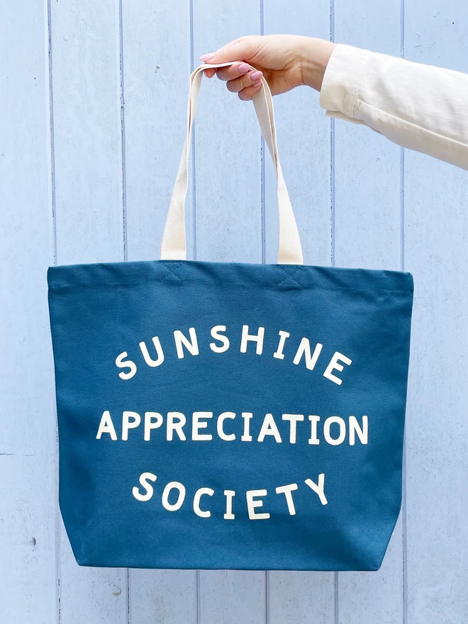 model holding out a large blue canvas tote bag with the words sunshine appreciation society on