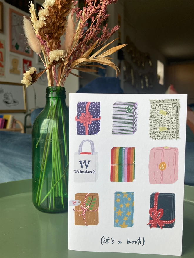Greetings card illustrated with books wrapped in various gift papers. Placed on a table with flowers in the background