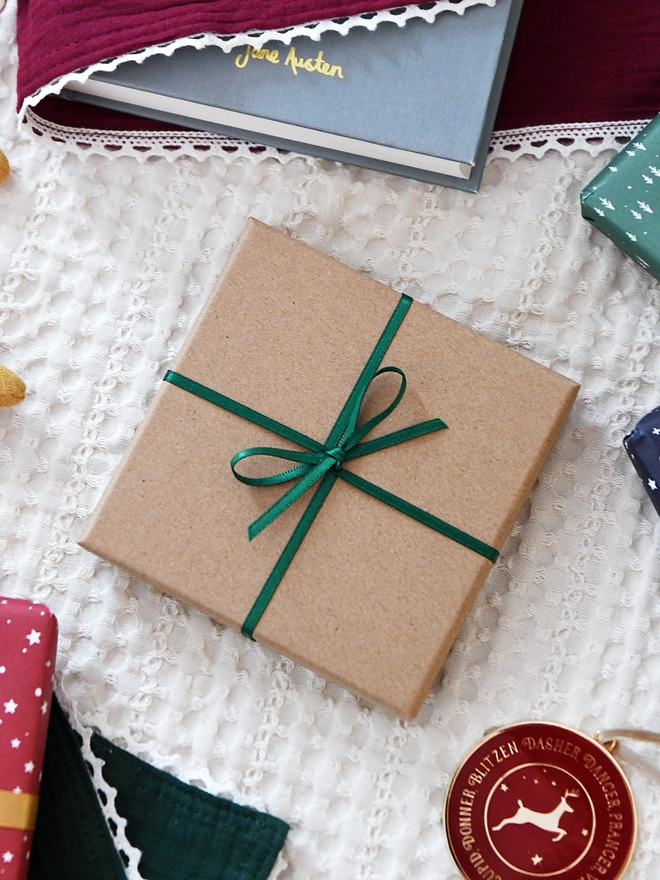A square kraft coloured gift box tied with green ribbon lays on white fabric and is surrounded by various wrapped gifts.