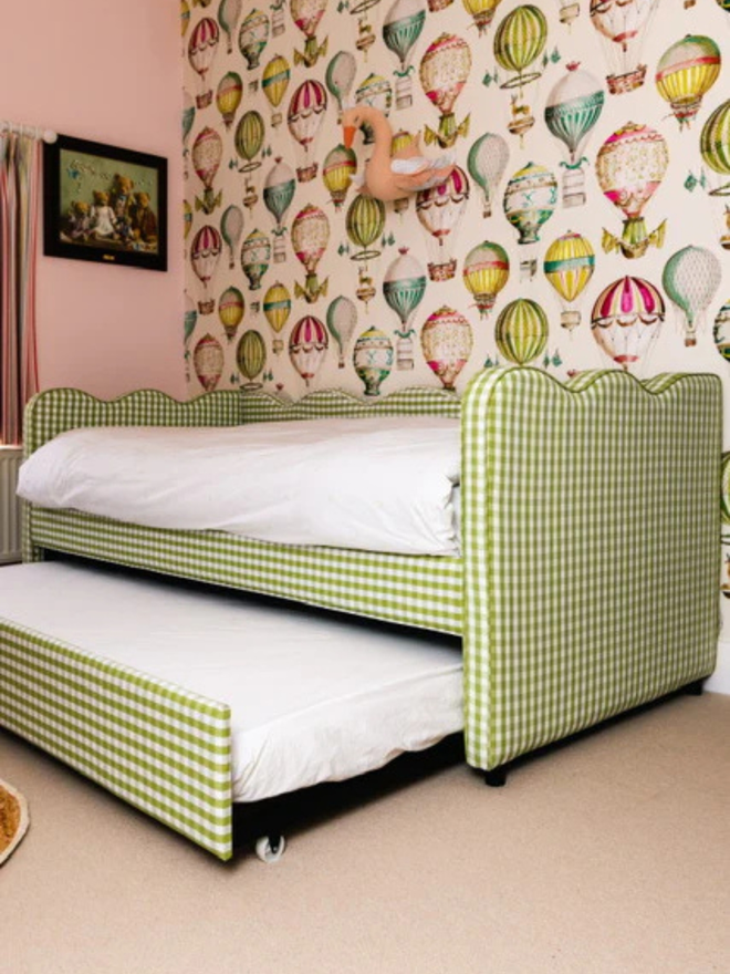 'the lochlan' green gingham upholstered children's bed. a green gingham sofa-style with a wavy headboard, with the trundle bed pulled out in a room with balloon-patterned wallpaper.