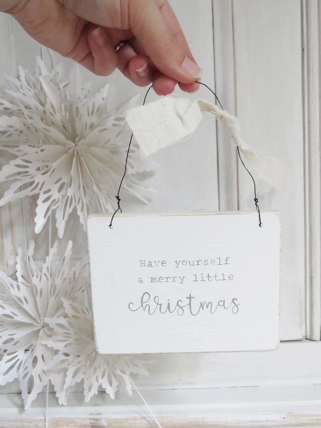 white wooden Christmas sign painted with a merry little Christmas message hung on wire and decorated with ribbon