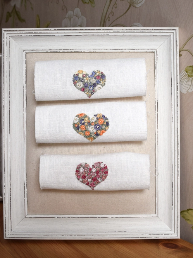A display of 3 Heart embroidery kits pinned to a decorative framed noticeboard, top to bottom are 1. Floral Meadow (Lavender Blues, Magenta & Buttermilk Yellow) 2. Sunshine Garden (Oranges, Yellows, Purples).  3. Pink Roses (5 shades of pink). 