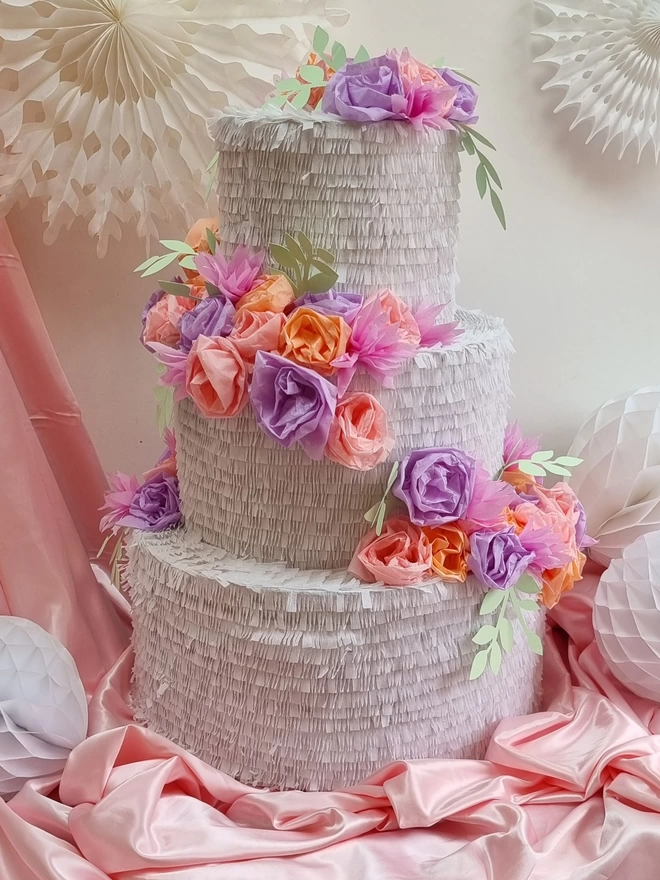 3 tier wedding cake pinata with a cascade of lilac and peach flowers and pale green leaves on a pink satin background with white paper fans