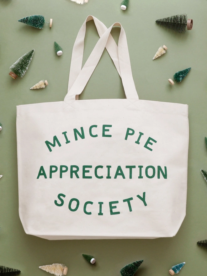 a large natural canvas tote bag bearing the words mince pie appreciation society laying on a green backdrop with mini christmas trees surrounding