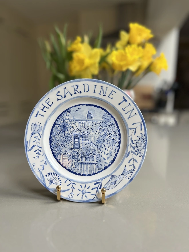A hand-painted blue and white plate stands with daffodils behind. It says “The Sardine Tin” in handwritten letters above with flowers around the rest of the rim, and inside a house with two dogs at the gate. 