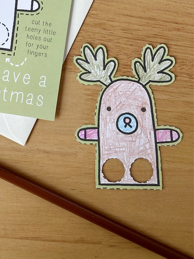 A colour in Christmas card with a reindeer finger puppet design lays on a white envelope on a wooden desk.