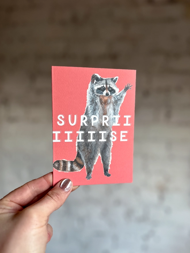 a bright coral pink greetings card featuring an illustration of a raccoon with its hands in the air with the text “surpriiiiise”