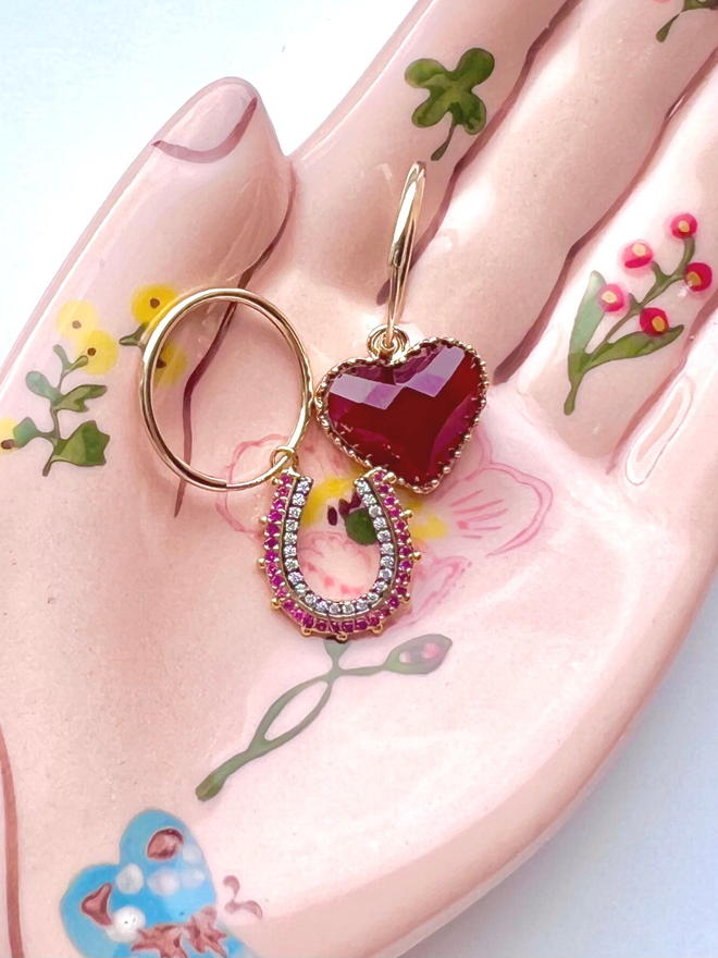 Small gold hoops threaded with a pink and white stone horseshoe charm and a red quartz heart charm, resting on a hand of hamsa plate