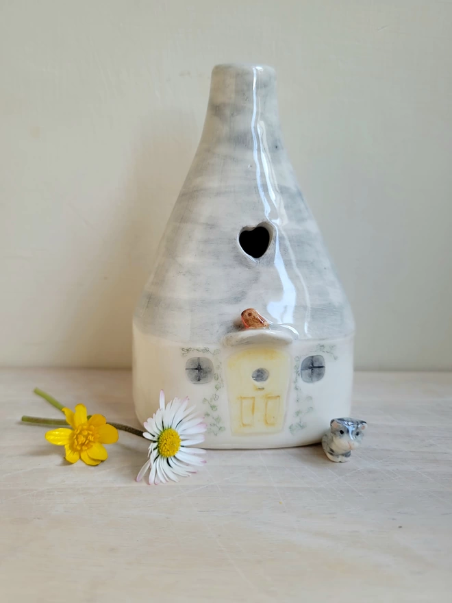 ceramic pottery house bud vase in the shape of a home with windows and yellow door above the door is a small robin bird and there is a cut out heart shape in the roof there is a daisy and butter cup lying on the light wood counter next to it   