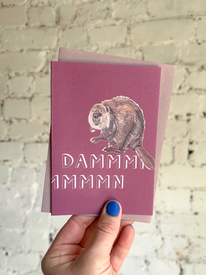 a pinky purple greetings card featuring an illustration of a beaver with the text “dammmmmn” with a muted purple envelope