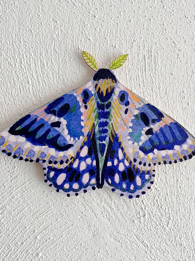 wooden moth wall decoration. blue wooden moth decoration hung on plain white wall.