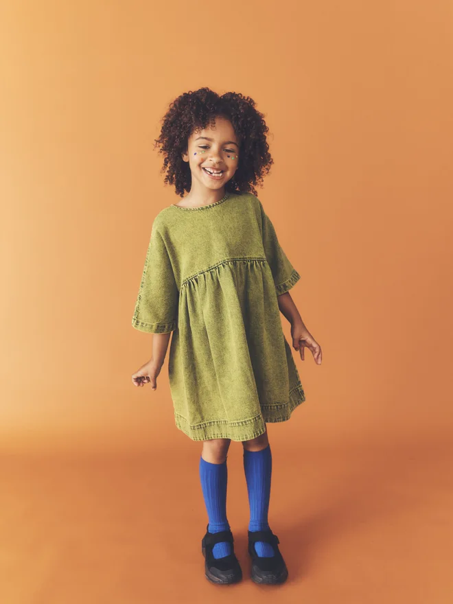 Child smiles against a burnt orange backdrop, wearing the Studio Koter Rumi dress in green acid-wash denim under a black bomber jacket, with cobalt knee socks.
