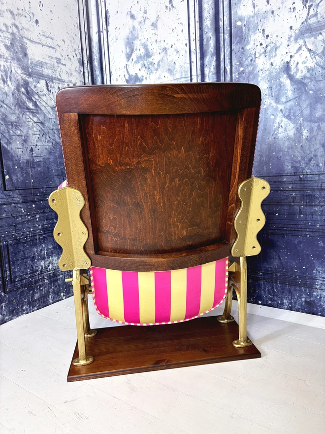 Rear view of a pink and yellow vintage cinema seat.  The cinema seat has gold legs and is mounted onto a wooden plinth.  The fabric is pink and yellow striped.