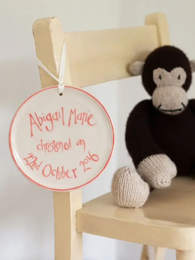 personalised ceramic birth commemoration plaque. plaque personalised with blue writing and hung on the side of a dining chair alongside a toy monkey.