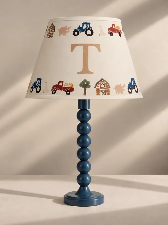 Personalised children's tractor lampshade by Studio Zef, displayed on a glossy navy blue bobbin table lamp against a warm neutral background. The cream empire shade features a large beige initial T, framed by hand-printed illustrations of blue tractors, red farm trucks, pink pigs, geese, barns and trees in earthy countryside colours.