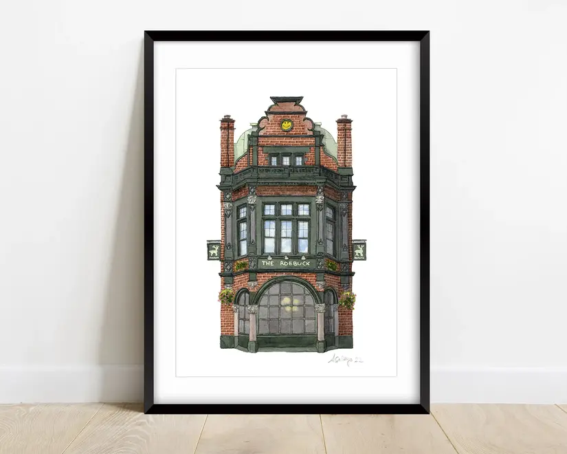 Borough The Roebuck Pub Giclée Print (Unframed), a print of a pub sitting in a black frame on a wooden surface against a white backdrop.