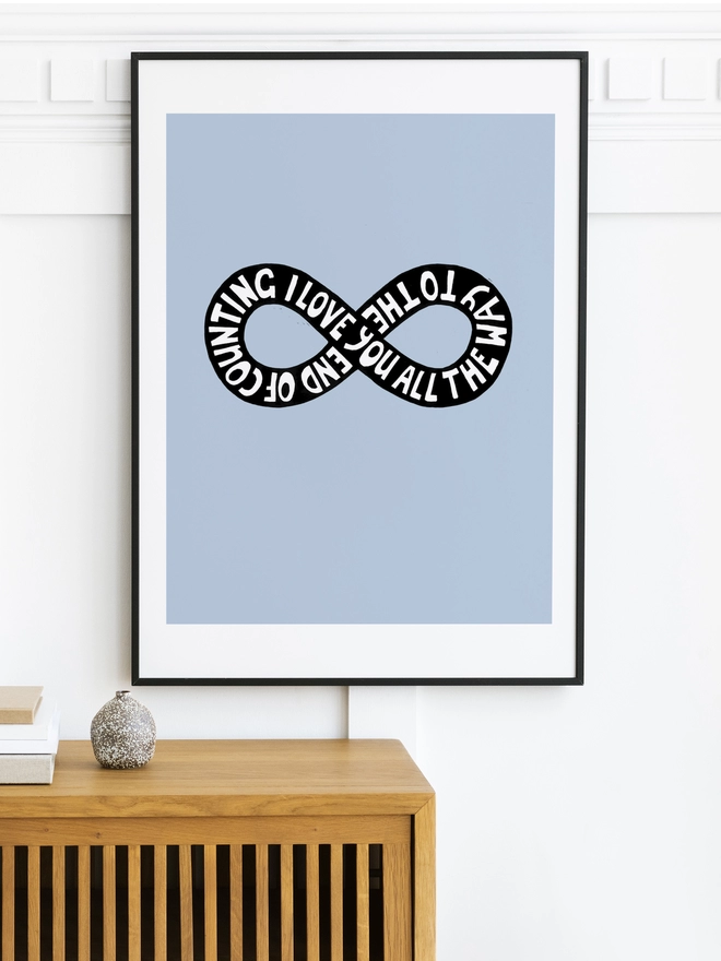 A pale blue framed print on a wall in a stylish home. The print has a black infinity design in black with white type going around it which reads: I Love You All The Way To The End Of Counting.