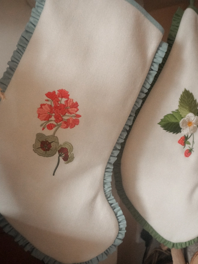 Two ivory embroidered Christmas stockings featuring geranium and strawberry botanical designs, finished with blue and green ruffled trims.