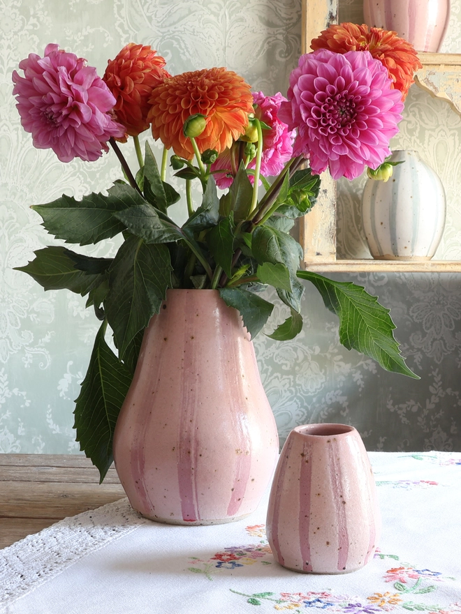 Handmade Ceramic Stoneware Small Vase in Pink on a wooden table with a vintage tablecloth. Behind the vase sits a large vase in the same style filled with orange and pink flowers.  