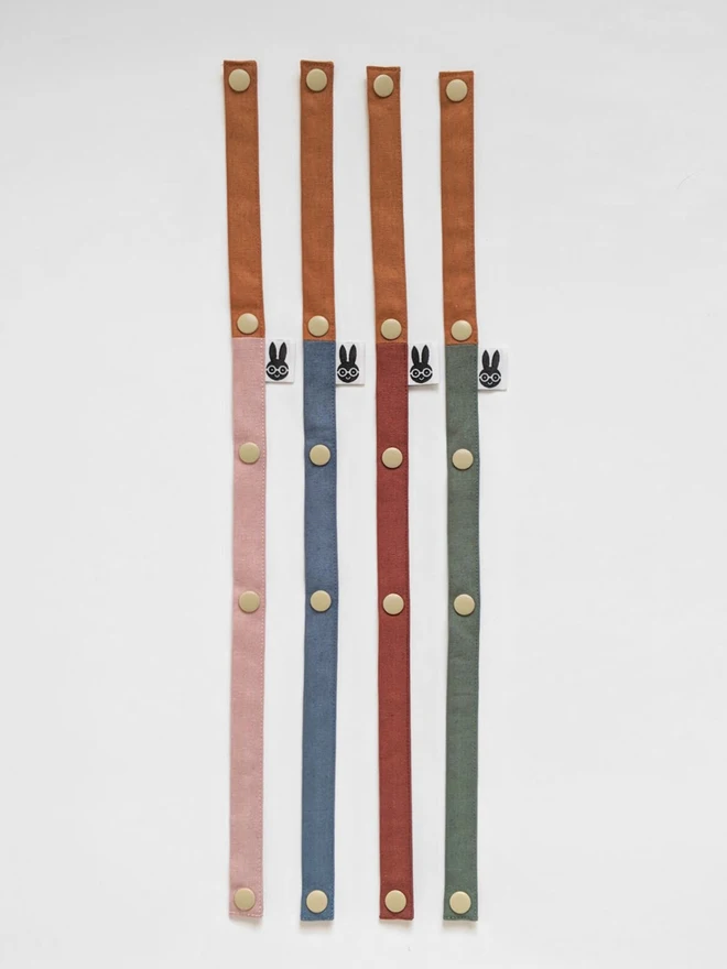 Handmade toy straps in woodland shades, featuring sturdy snap fastenings and small fabric tags with a bunny logo, neatly arranged in a row.