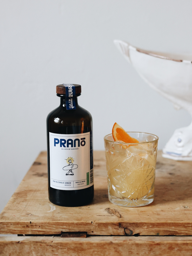 Bottle of prano sitting next to a glass of the pale orange beverage garnished with a slice of orange. Both sitting on a brown countertop with white background 