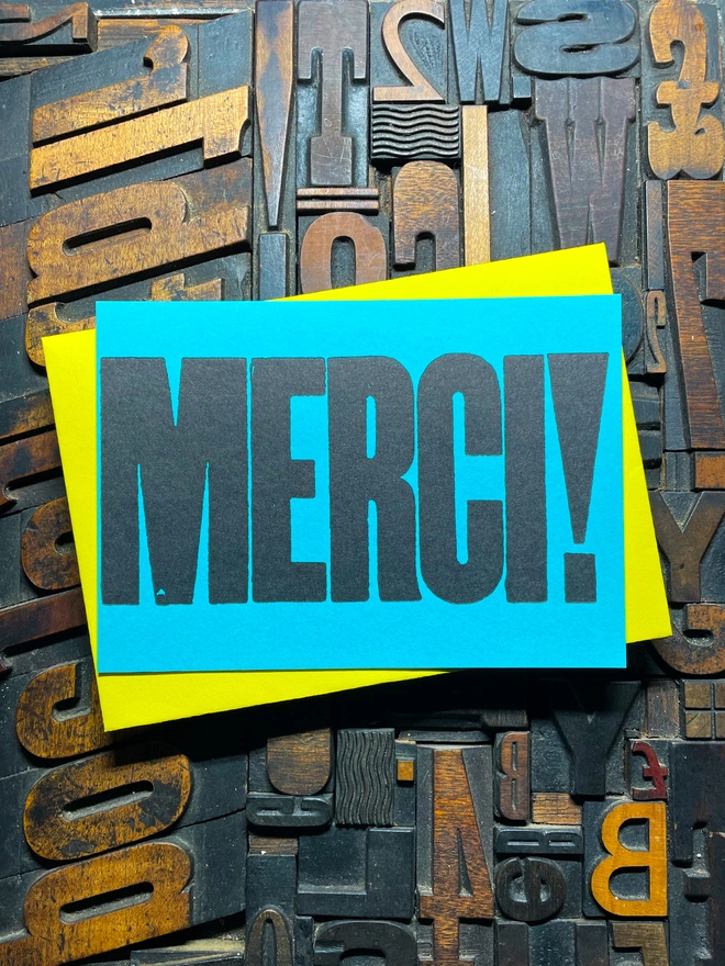 A bright turquoise thick duplex card with the word "MERCI!" printed in bold, black letters lies on a surface covered with various letterpress blocks.