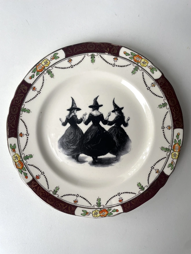 a coven of 3 witches dance hand in hand, the image is victorian in black and white and is printed directly onto an ornate vintage plate