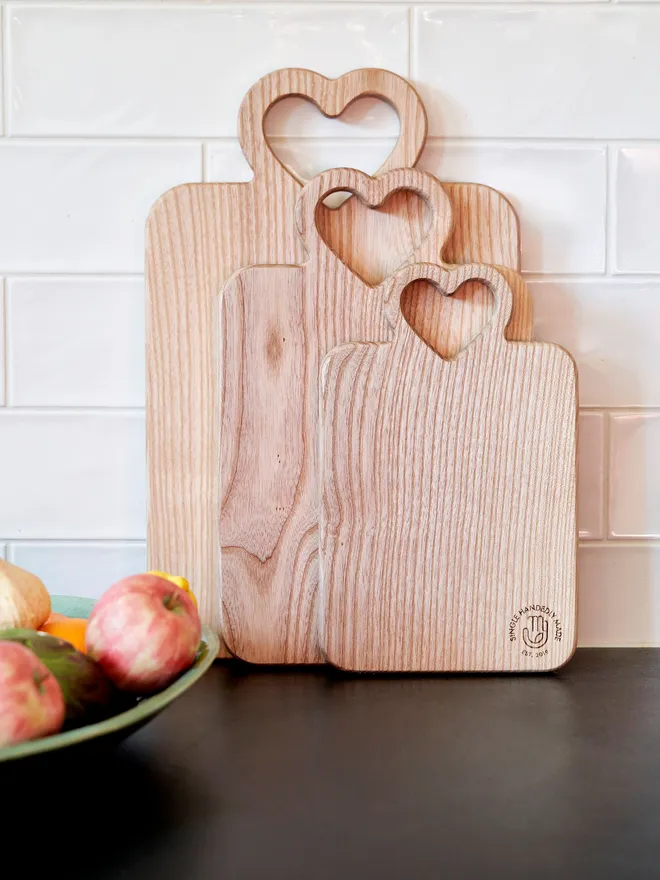 Set of 3 heart wooden serving boards leaning against a white tiled wall with black worktop and a fruit bowl