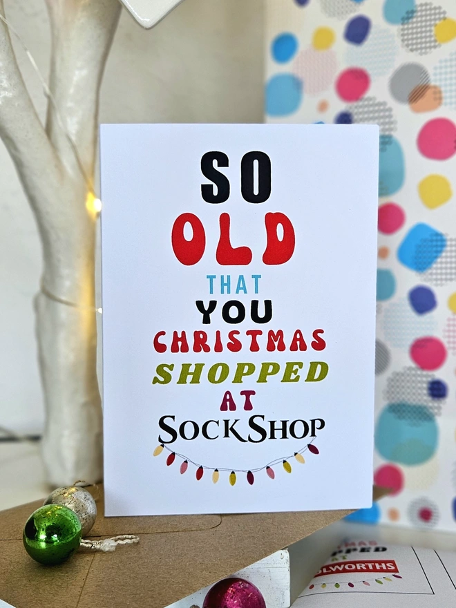 A photograph of the front of a humorous A6 greeting card illustrated using brightly coloured text and the retro shop logo. The text says so old  that you Christmas shopped at Sock Shop with string lights