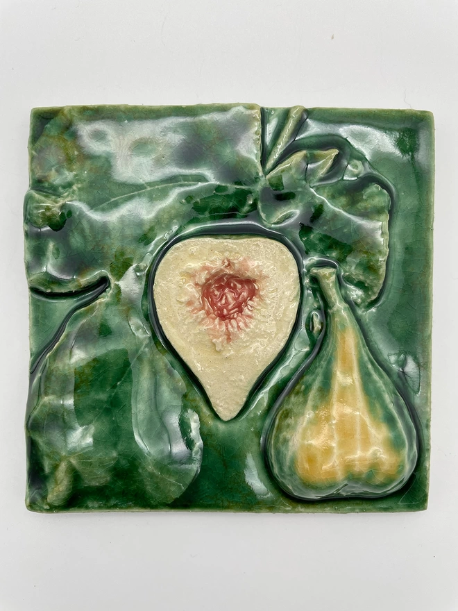 Handmade ceramic tile taken from a plaster cast of a real fig, cut open to show one half fleshy side up and one half skin side up, plus fig leaves. Very realistic, three-dimensional, with lush coloured glazes.
