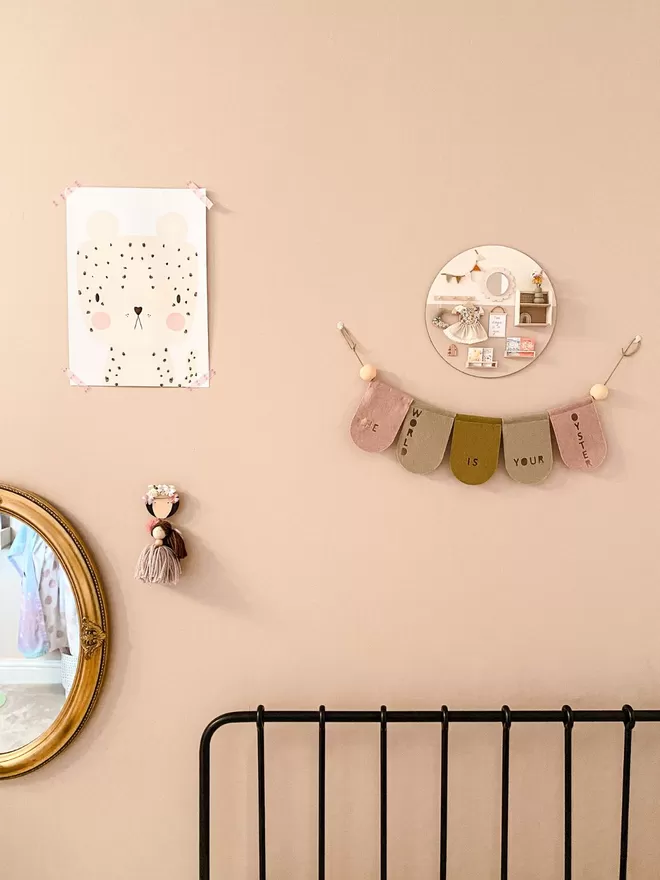 Garland hanging on pale pink wall, bed frame in view, half a mirror and a small circular plaque hanging.