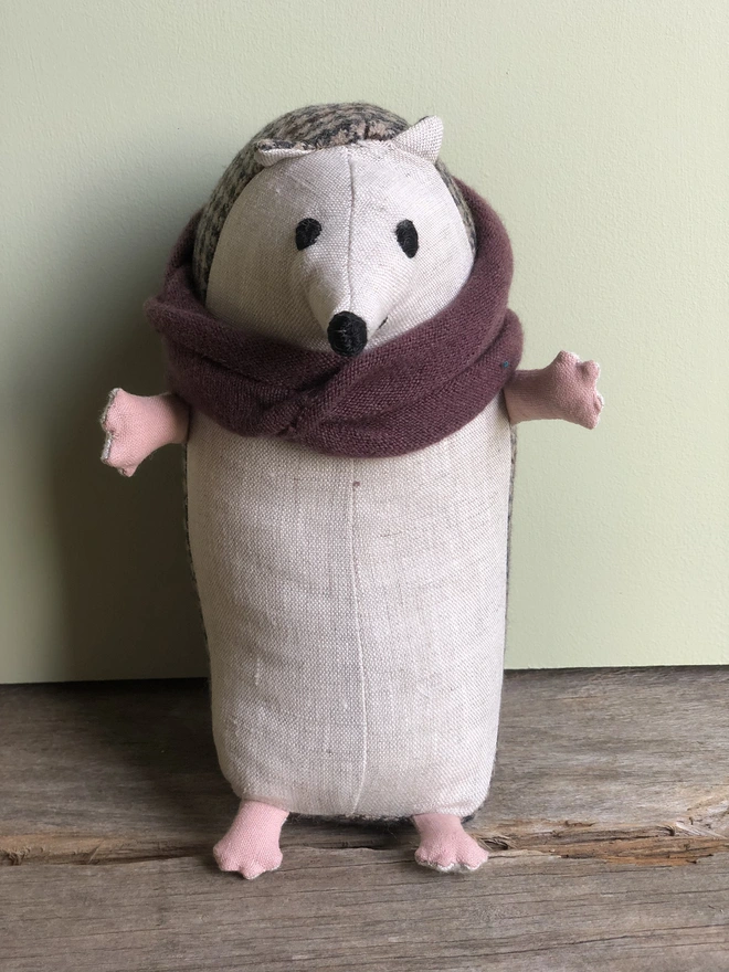 Handmade toy hedgehog, made from natural linen (front) and brown tweed (back). Wearing a burgundy lambs wool scarf with his arms open. 