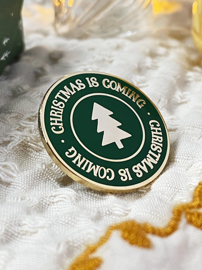 A deep green round enamel pin badge with a gold Christmas tree in the centre and gold lettering around the outside that reads "Christmas Is Coming" rests an ivory fabric.