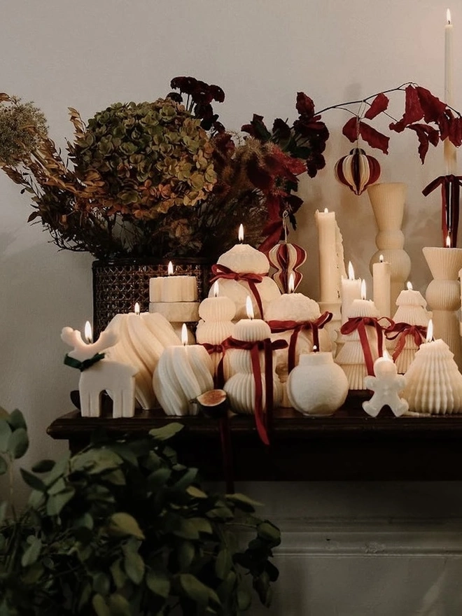 Reindeer Sculpture Candle. A mantelpiece display of cream festive candles, including a reindeer, gingerbread man, Christmas trees and swirled pillars tied with red velvet ribbons, styled with dried hydrangeas and autumn foliage. The elegant handmade candles bring warmth and seasonal charm to holiday décor.