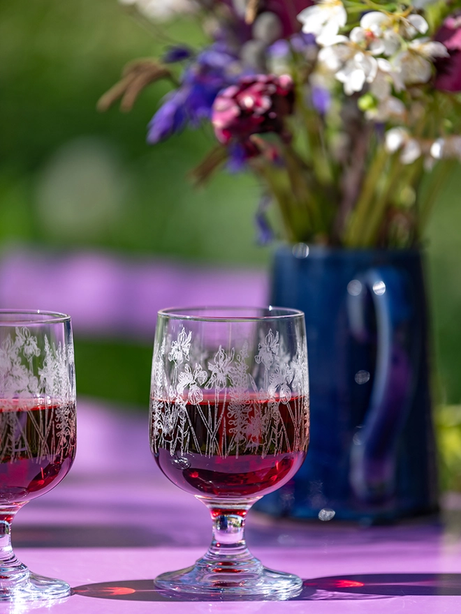 A short-stemmed bistro glass decorated with, illustrated poppies, hydrangeas, and sunflowers against a garden background.