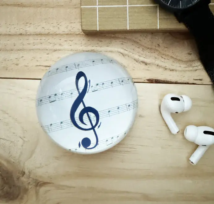Round paperweight with a treble clef and sheet music on a wooden surface beside earbuds.