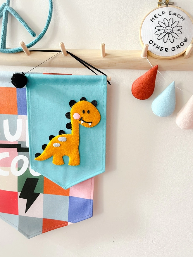 A mustard coloured dinosaur on a blue canvas fabric banner hung up on a peg rail