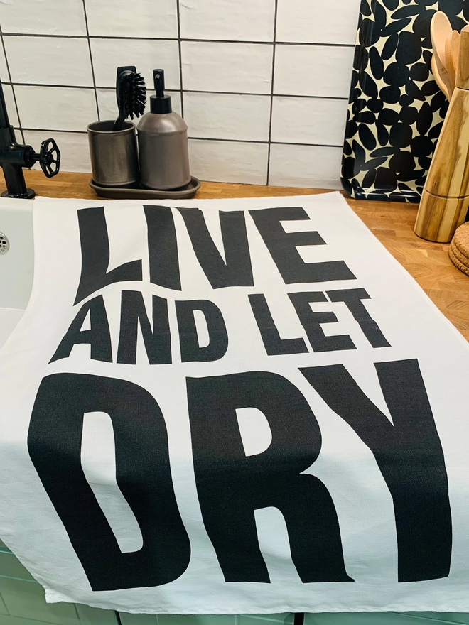 London Drying Live and Let Dry black print on white tea towel laying on kitchen counter 