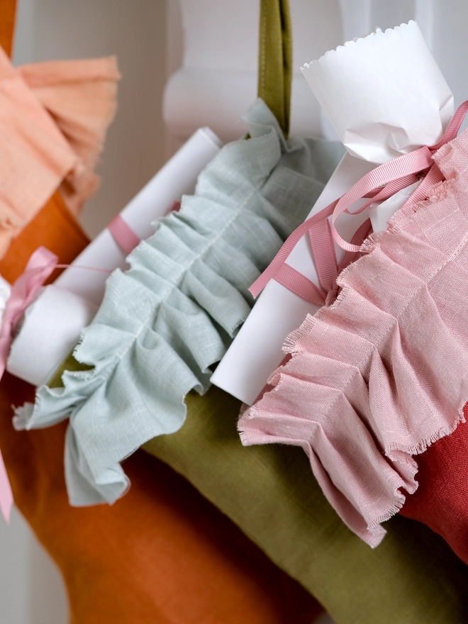 handmade linen Christmas stockings in warm orange, olive green and red hang beneath a festive mantel, each finished with a soft, gathered ruffle cuff.