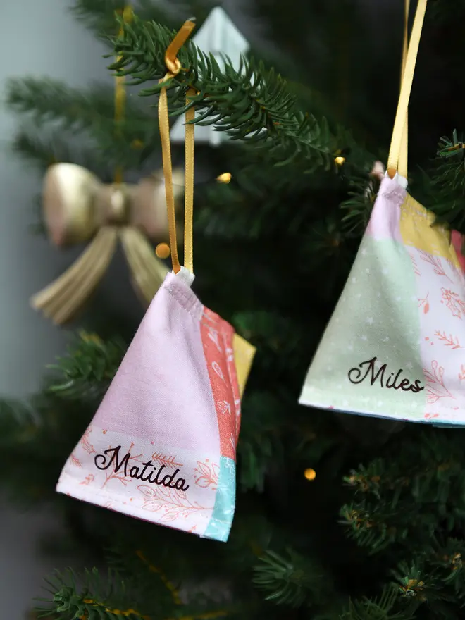 A handmade fabric fortune cookie Christmas decoration in pastel patchwork print, personalised with the name “Matilda” in brown, hangs from a golden ribbon on a Christmas tree branch.