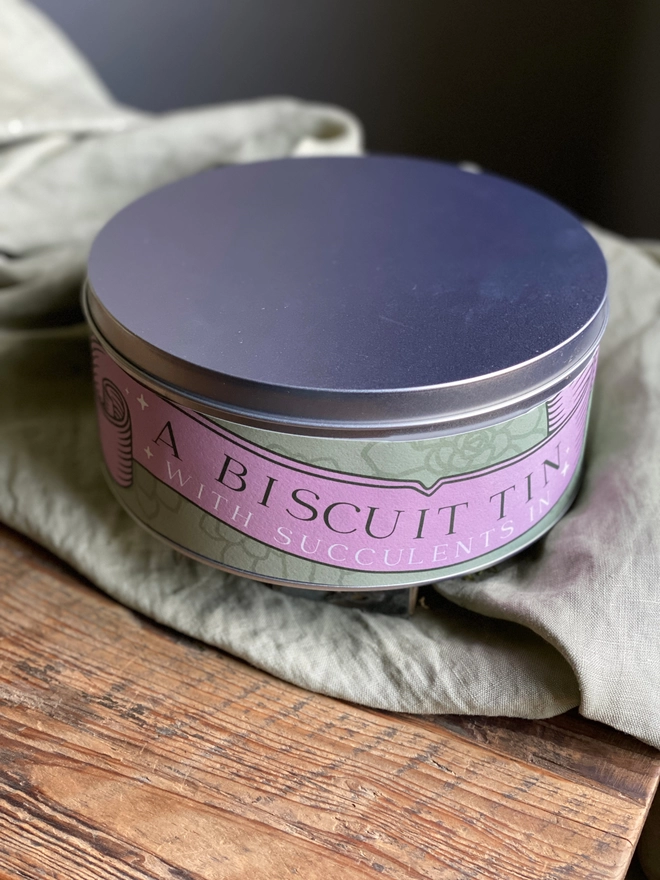 A closed silver biscuit tin. The label on the tin reads 'A Biscuit Tin With Succulents In' on an illustrated pink banner. 