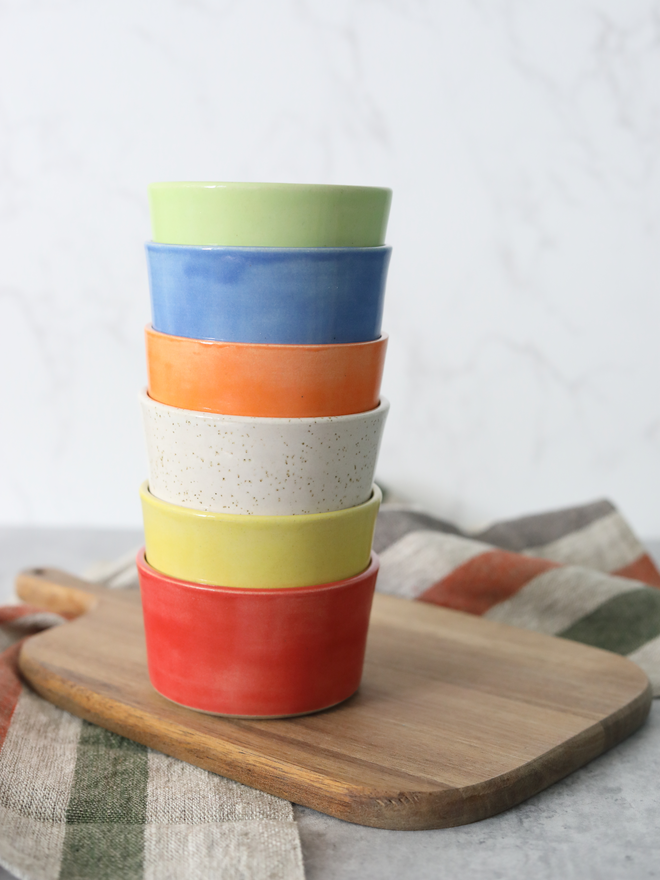 Six handmade ceramic side bowls stacked in a tall tower on a wooden board, showing a colour stripe of spring green, blue, mandarin orange, speckled white, yellow, and cherry red, styled with a multicoloured striped linen cloth against a white marble background
