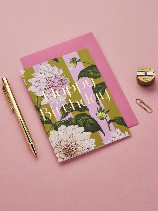 happy birthday card with gold foil wording and illustrated Dahlia flower on a green and pink stripes base and a hot pink envelope