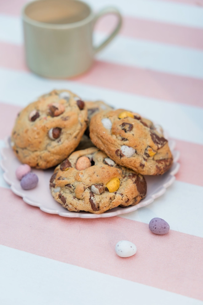 Chunky Mini Egg cookies on a scalloped plate atop a pink and white striped tablecloth - a mug just out of shot and scattered Mini Eggs
