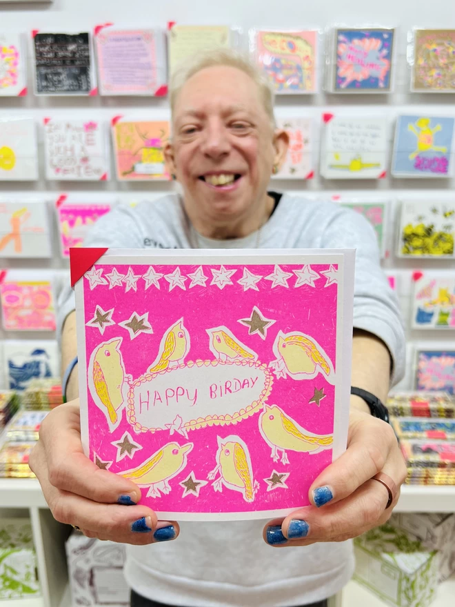 Artist holding a riso printed birthday card featuring birds in yellow & pink with the words Happy Birday