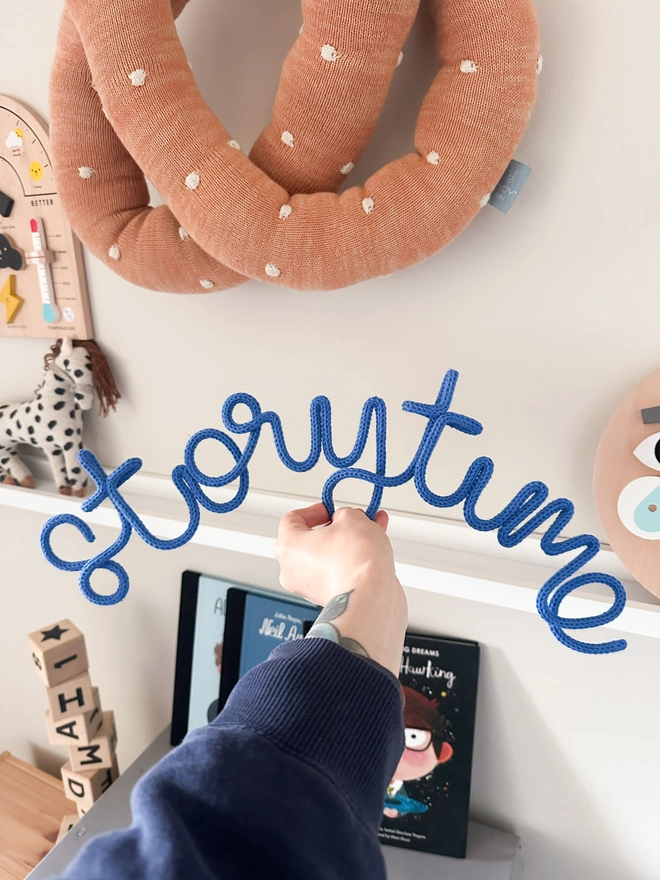 A storytime knitted wire word sign being held up infront of a childrens reading area with kids books and decor. 