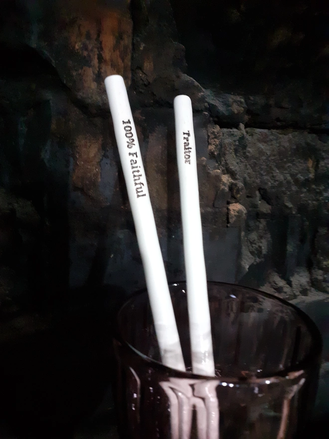 Handmade ceramic drinking straws inspired by The Traitors TV show — one inscribed ‘Traitor’ and the other ‘100% Faithful,’ finished in glossy white glaze with black lettering. The pair stand in a smoky brown glass against a rustic brick wall, softly lit to evoke the show’s moody atmosphere.